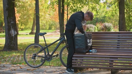 bicycle courier man inspects delivery bag on bench, unloading takeout containers in autumn park with leafy path and bicycle leaned nearby, focused expression while checking order on smartphone - Powered by Adobe