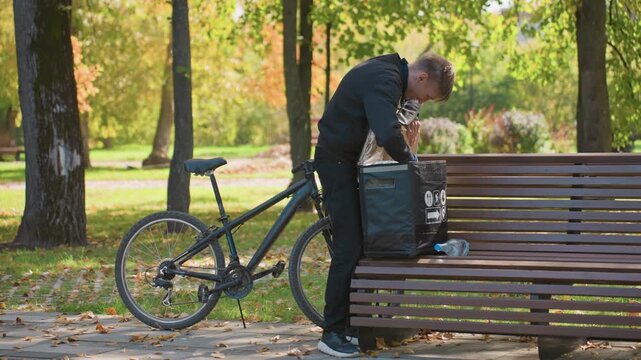 bicycle courier man inspects delivery bag on bench, unloading takeout containers in autumn park with leafy path and bicycle leaned nearby, focused expression while checking order on smartphone