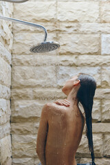 Woman taking outdoor shower with water flowing down back, natural stone background, wet hair, relaxing moment, warm light, summer wellness.