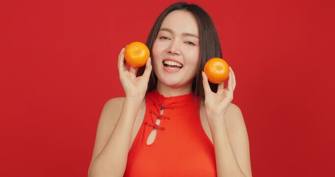 Happy young Asian woman in red Chinese dress holding two oranges on red background for Chinese New Year celebration and luck concept.