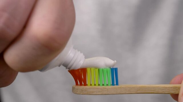 Hand holds bamboo toothbrush with rainbow bristles while applying white toothpaste. Concept of eco routine, mindful habits, sustainable hygiene, and morning freshness