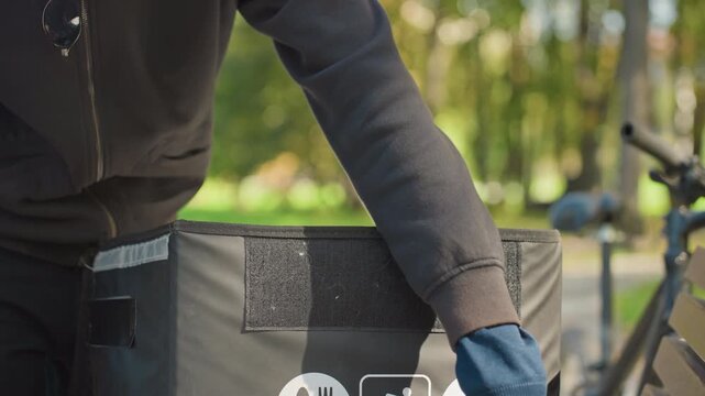 white courier inspects insulated food bag, outdoor park beside parked bicycle, hands unzip thermal foil liner to check orders, adjusts seal and prepares contactless takeaway handoff, casual hoodie