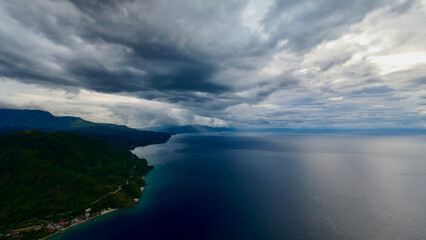 Dramatic aerial view of tropical coastline under heavy storm clouds. Dark ocean water meets green mountains, creating a moody seascape with approaching weather.