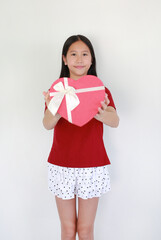 A charming young Asian girl in a red t-shirt and polka dot skirt smiling while holding a heart-shaped gift box with a white ribbon, standing against white background.