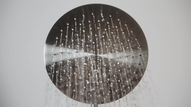 Close-up looking up shot round stainless steel rainfall shower headon white background in bathroom as water begins to flow and stops. Thin streams emerge from holes creating cascade in slow motion