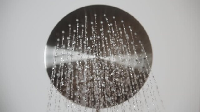 Close-up looking up shot of round stainless steel rainfall shower headon white background in bathroom as water begins to flow. Thin streams emerge from holes creating cascade in slow motion