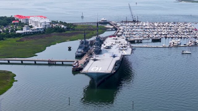 Aerial drone footage showing a USS Yorktown Aircraft Carrier docked at a harbor, with a long boardwalk and surrounding marina in the background
