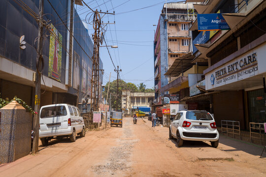 Kannur, India - March 13, 2023: A dirt street in Kannur shows parked cars, an auto rickshaw, pedestrians, retail shops with signage in English, and visible power lines under clear daylight.