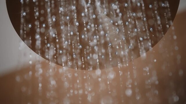 Close-up of modern rainfall shower head as water gradually turns on. Round stainless steel shower head with matte metallic finish. Curved arm, warm brown ceramic tiles, water flow starts and stops