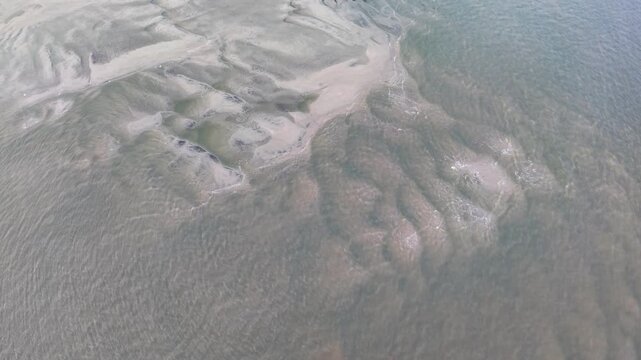 Drone footage capturing patterns of sand and shallow water along beach, with swirling sediment and calm waters creating unique textures