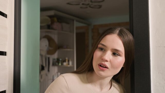 young caucasian woman opens apartment door and peers through peephole, smiling as she checks for delivery cozy hallway with shelves and soft lighting, neutral sweater, subtle head tilt, leaning