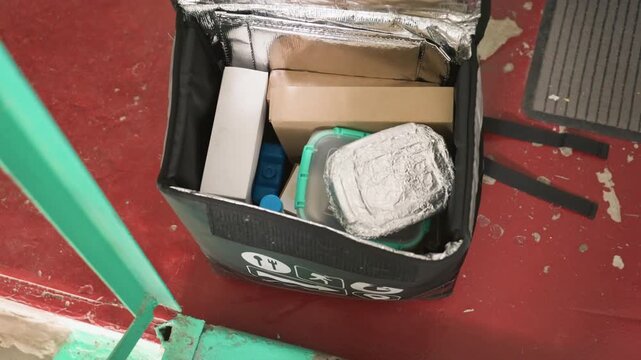 Courier unpacking insulated food bag, hands unzip thermal liner revealing foil containers and cardboard boxes, checking seals and arranging meals for delivery. Red bus floor and green rail form urban