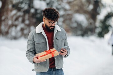 Obraz premium Man with curly hair wearing a gray jacket and red sweater is holding a gift box with a ribbon while checking his phone outdoors in a snowy environment with trees in the background