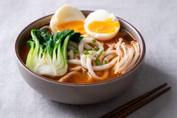Spicy Udon Noodle Soup topped with Bok Choy, Enoki Mushrooms and Soft Boiled Egg in a bowl with chopsticks on a fabric background