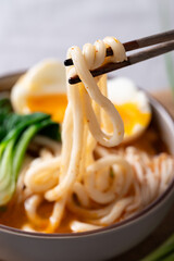 Close up of chopsticks picking up spicy udon noodles from a bowl with bok choy, enoki mushrooms and soft boiled egg