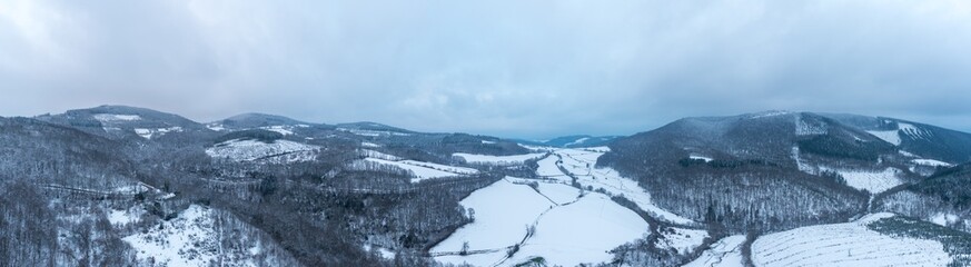 Expansive aerial panorama of snow-blanketed valleys and forested hills in the Morvan region. Soft winter light and a cloudy sky create a peaceful, remote atmosphere.