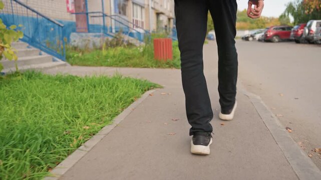 white courier walking with insulated backpack along suburban sidewalk past parked cars and patchy grass, approaching residential doorstep while navigating pavement, reflective strip on bag, casual