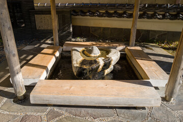 A rustic stone hot spring foot bath with wooden benches is sheltered under a simple pavilion in the center of Kinosaki. Sunlight highlights the textures of weathered wood, natural stone, and flowing