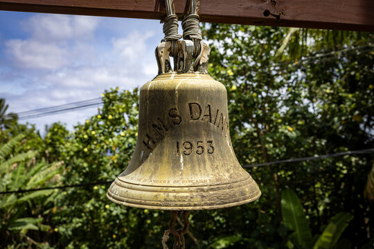 Adamstown, Pitcairn Islands - 02 January 2026: View of the aged, weathered bell, 'H.M.S. Dainty 1953', suspended against a backdrop of lush, vibrant greenery and a soft, diffused sky.