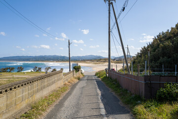 Obraz premium A quiet paved lane bordered by fences and utility poles descends toward the sandy shore of Shotenkyo Beach in Kumihamacho, with blue waves and distant mountains under a clear sky.