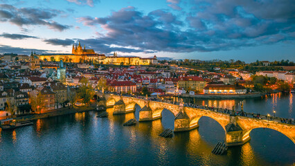 Prague Castle and Charles Bridge are illuminated at night. This aerial view shows the Vltava River and the city of Prague during evening hours. The lights reflect on the water. © pyty