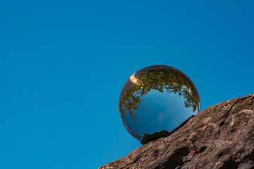 A clear crystal ball resting on a rock captures an inverted reflection of trees and blue sky, creating a surreal and artistic nature scene. This image conveys concepts of perspective reflection