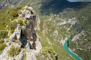 Jagged limestone cliffs rise sharply above a vivid turquoise river snaking through a deep, forested canyon in the Mediano area of Aragon, Spain. Bright sunlight accentuates the rugged textures and