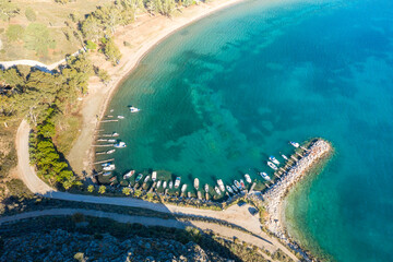 Fototapeta premium Aerial view of a curved bay with a small marina and boats docked along a rocky breakwater at Karathona Beach near Nafplio, Greece. The clear turquoise water contrasts with the sandy shoreline and lush