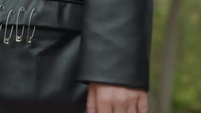 closeup leather jacket pocket safety pins, woman standing outdoors wearing black leather coat with metal pins clipped to pocket, green sweater peeking at waist, hand relaxed by side, soft forest