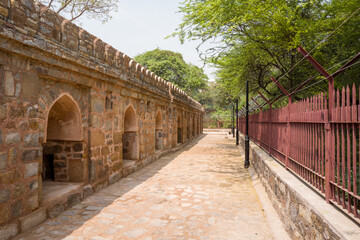 Obraz premium Sunlit stone arcade with arched niches lines a cobblestone walkway in Mehrauli Archaeological Park, Delhi. The historic wall contrasts with iron fencing and dense green trees under a clear sky.