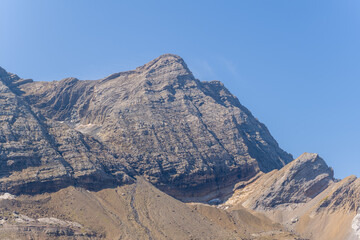 Imposing mountain peak with rugged stone layers rises sharply against a cloudless blue sky in the Pyrenees near Gavarnie Gedre. Sunlight highlights the textured rock surfaces and dramatic geological