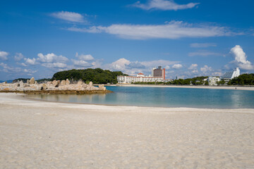 Obraz premium A stone jetty extends into calm blue water beside a wide stretch of white sand, with modern hotels and lush green trees under a bright sky in Shirarahama, Japan.