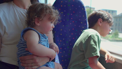 Obraz premium Baby sitting upright in mother’s arms looking out on bus while older child leans against window, both watching passing city scenery