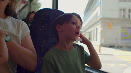 Mother lifting blouse to breastfeed baby while sitting on bus, older sibling beside them looking...