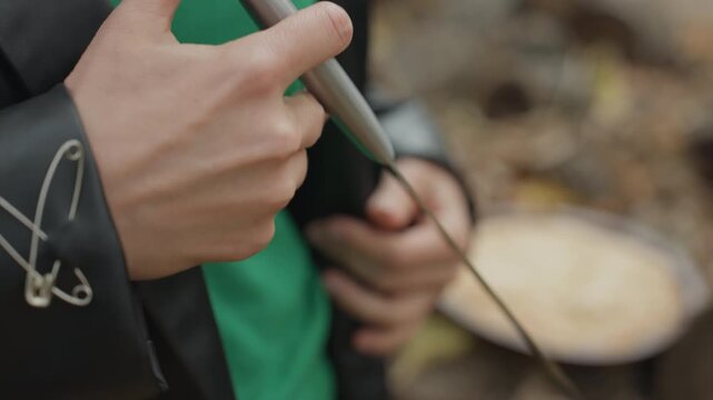 hands sharpening knife beside pie on tree stump, leather jacket with safety pins, green sweater, metal file honing blade, pastry resting on wood, autumn leaves scattered, intimate closeup, tactile