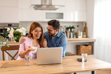 Obraz premium Happy Young Couple Looking at Laptop Screen Together in Modern Kitchen. Cheerful Man and Woman Discussing Online Shopping or Good News at Home. Lifestyle and Technology Concept.