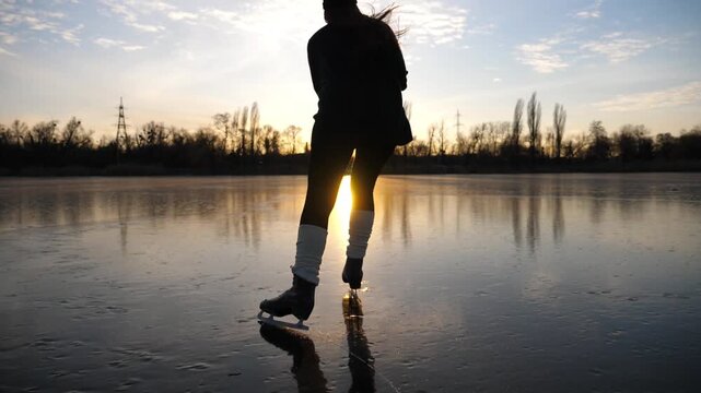 Female skater shod in figure skates sliding on ice surface during beautiful sunset. Sportswoman skating on frozen river. Silhouette of woman training at lake. Shining sun reflecting on surface of ice