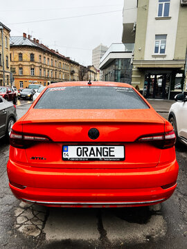 Bright orange Volkswagen Jetta parked on a wet city street with modern buildings in the background