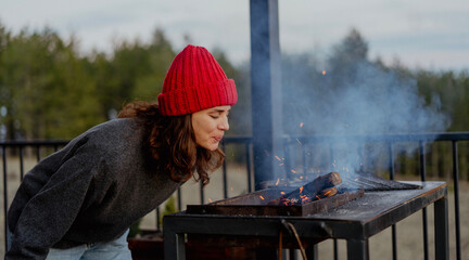 Young woman in red hat blowing fire in barbecue grill on countryside house terrace