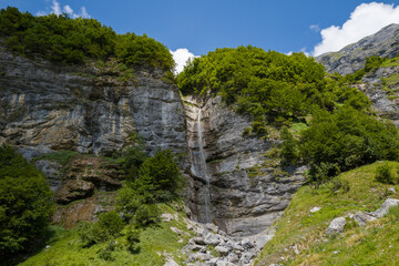A slender waterfall spills down a rugged limestone bluff surrounded by lush green trees and rocky slopes in Cirque du Fer-a-Cheval under a bright blue sky.