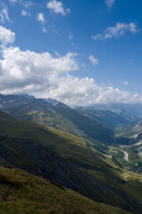Fototapeta premium Sweeping view of green mountain slopes and deep valley in Val Ferret, with dramatic cloud formations and bright blue sky creating a sense of depth and tranquility.