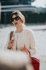 Stylish woman in a cream coat and oversized sunglasses gestures outdoors, holding a vape near a red bag.
