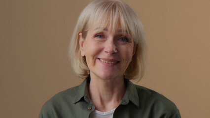 Emotional friendly Caucasian old woman look smiling looking at camera cheerful positive satisfied retired female senior lady 60s glad granny posing in beige studio smile with healthy teeth enjoying