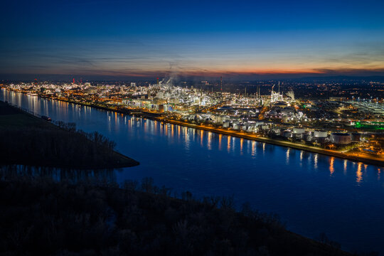 Aerial view of sprawling BASF industrial complex ablaze with golden lights reflecting on the dark waters of the river at twilight, Ludwigshafen am Rhein, Rheinland-Pfalz, Germany.