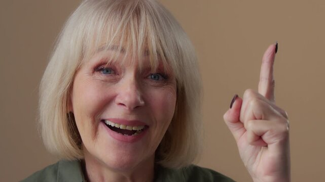 Portrait Caucasian old woman come up with idea finding answer happy smiling looking at camera in beige studio mature female senior retired granny raising index finger remind insight gesture solution