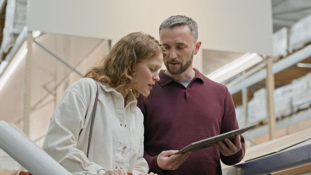 Low angle shot of smiling man holding sample of parquet while his wife chatting with him and touching parquet