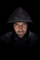 A DJ stands in a dark space wearing a hood. He looks directly at the viewer with a serious expression. Headphones rest around his neck as he prepares to perform.
