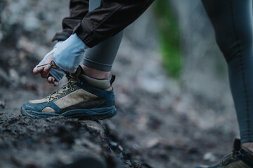A woman ties the laces of her hiking boot on a rocky trail. The scene shows outdoor activity and preparation for a trek in nature.