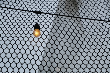 Single warm light bulb hanging in front of wire mesh fence with soft sky background