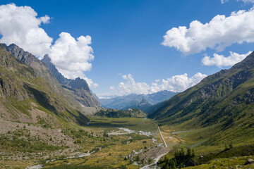 Fototapeta premium Expansive view of a green alpine valley flanked by steep rocky mountains under a bright blue sky with scattered white clouds near Col de la Seigne in the Italian Alps. Sunlight highlights the rugged